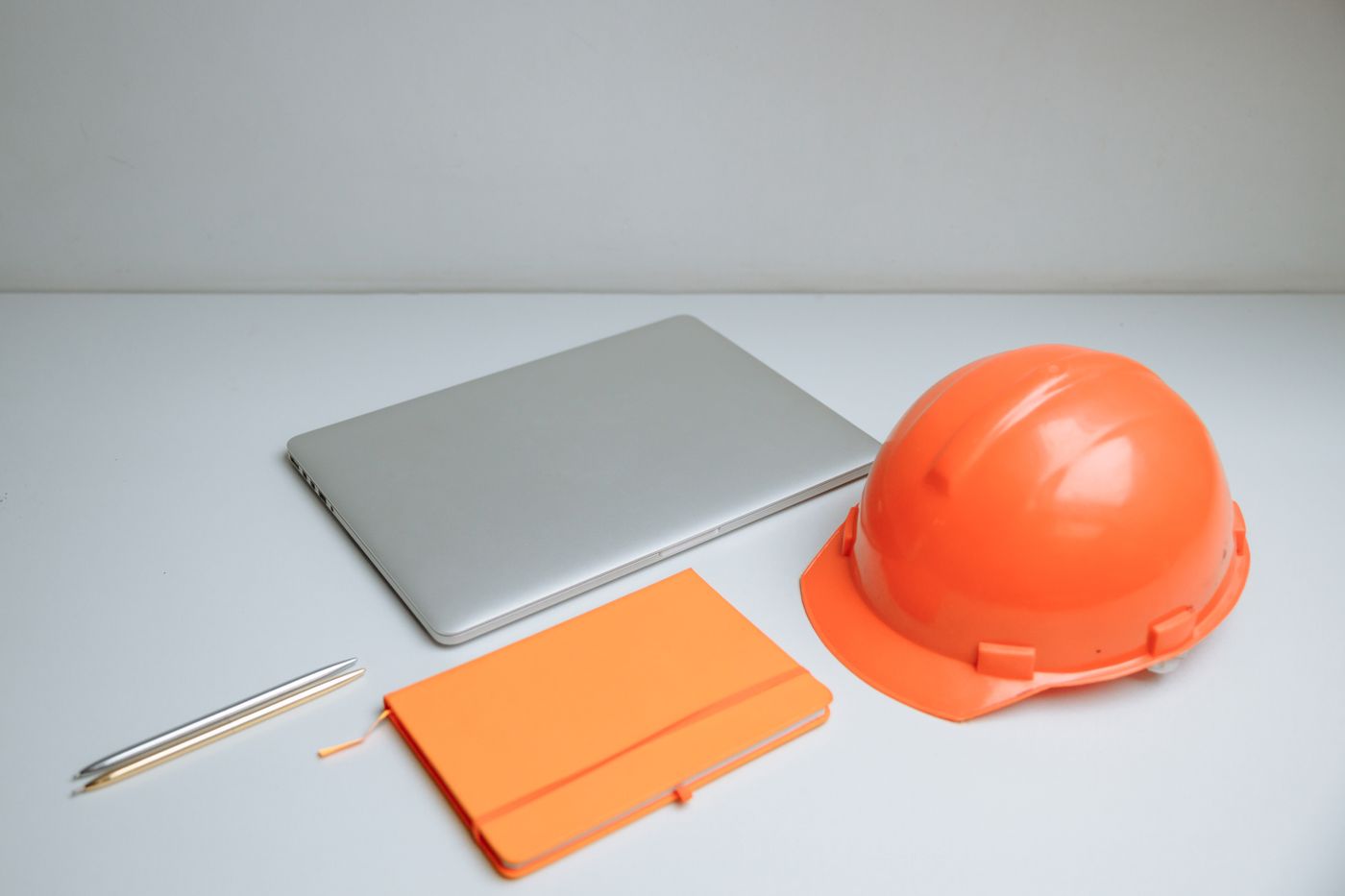 A laptop, a notebook and an orange hard hat on a plain desk.