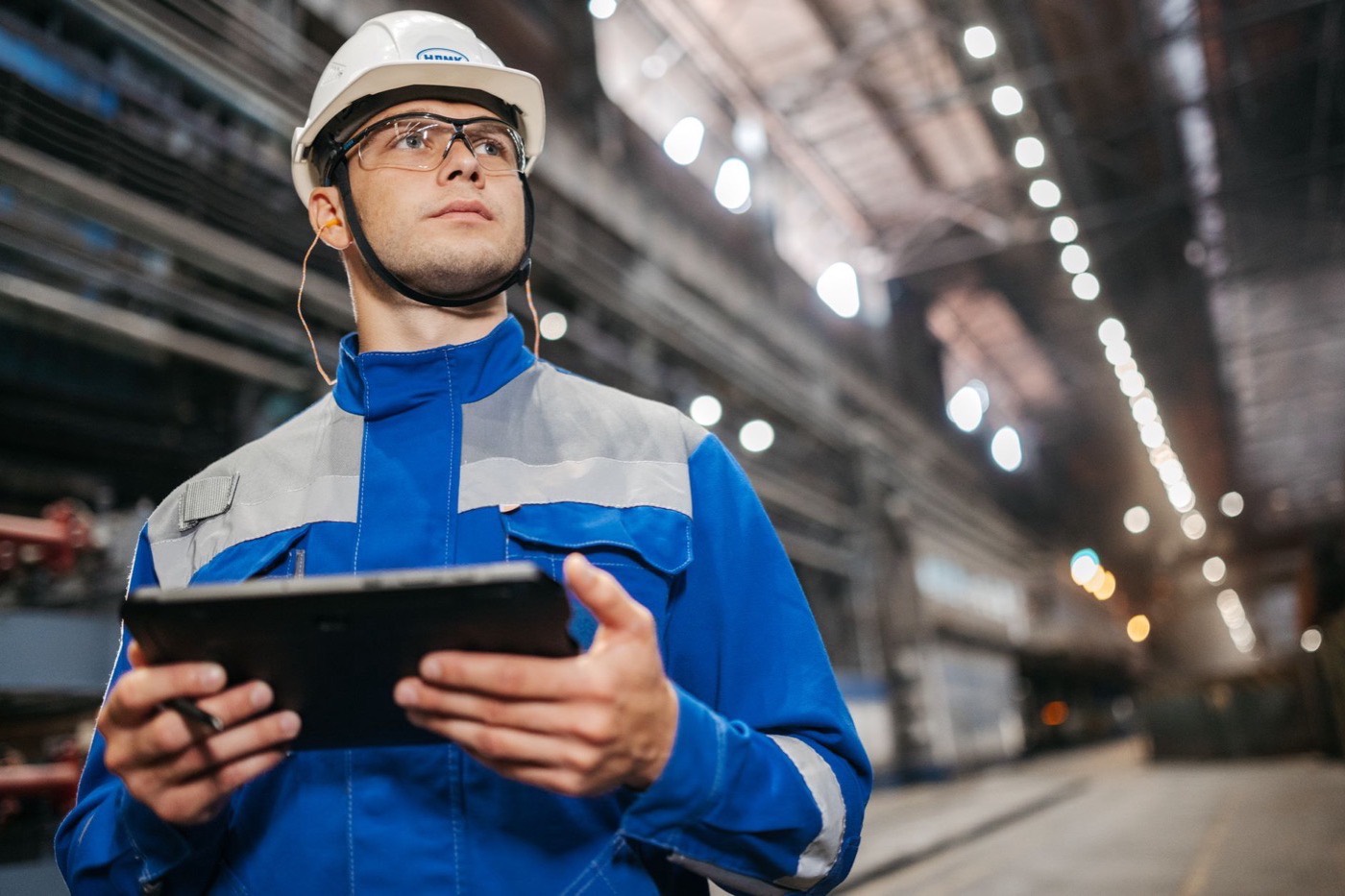 Factory worker in safety gear using a tablet in a well-lit industrial setting.